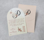 Προσκλητήριο γάμου με απαλό σομόν φάκελο και οβάλ κάρτα με εκτυπωμένα σχέδια και μονόγραμμα.